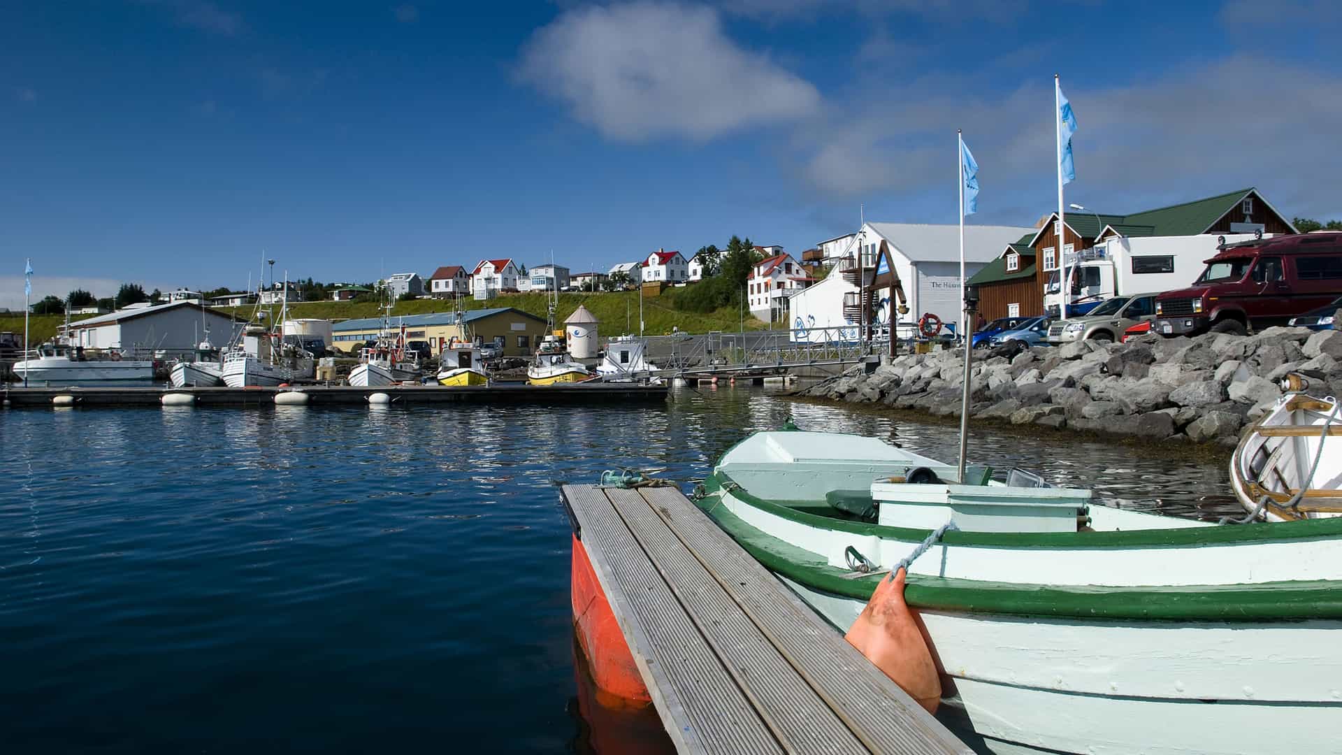 A beautiful view of the harbor in Husavik, Iceland, with fishing boats docked in the calm water, and a colorful town with red and white houses lining the hill in the background.