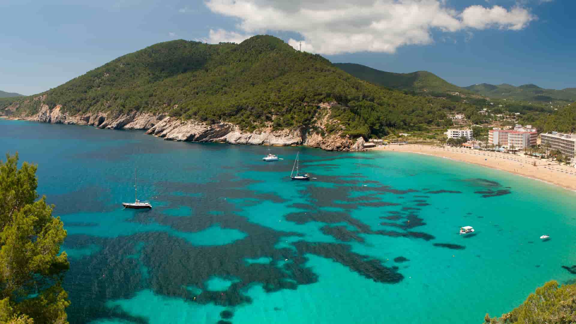 A scenic aerial view of a vibrant turquoise bay in Ibiza, Spain, with boats anchored in the clear water, a sandy beach, and lush green hills surrounding the coastline.