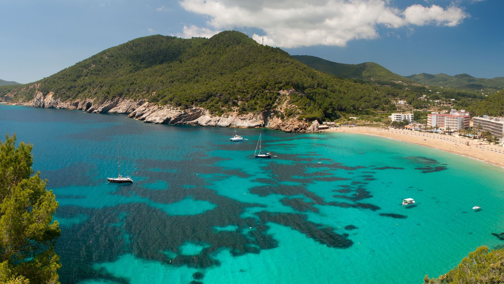 A scenic aerial view of a vibrant turquoise bay in Ibiza, Spain, with boats anchored in the clear water, a sandy beach, and lush green hills surrounding the coastline.