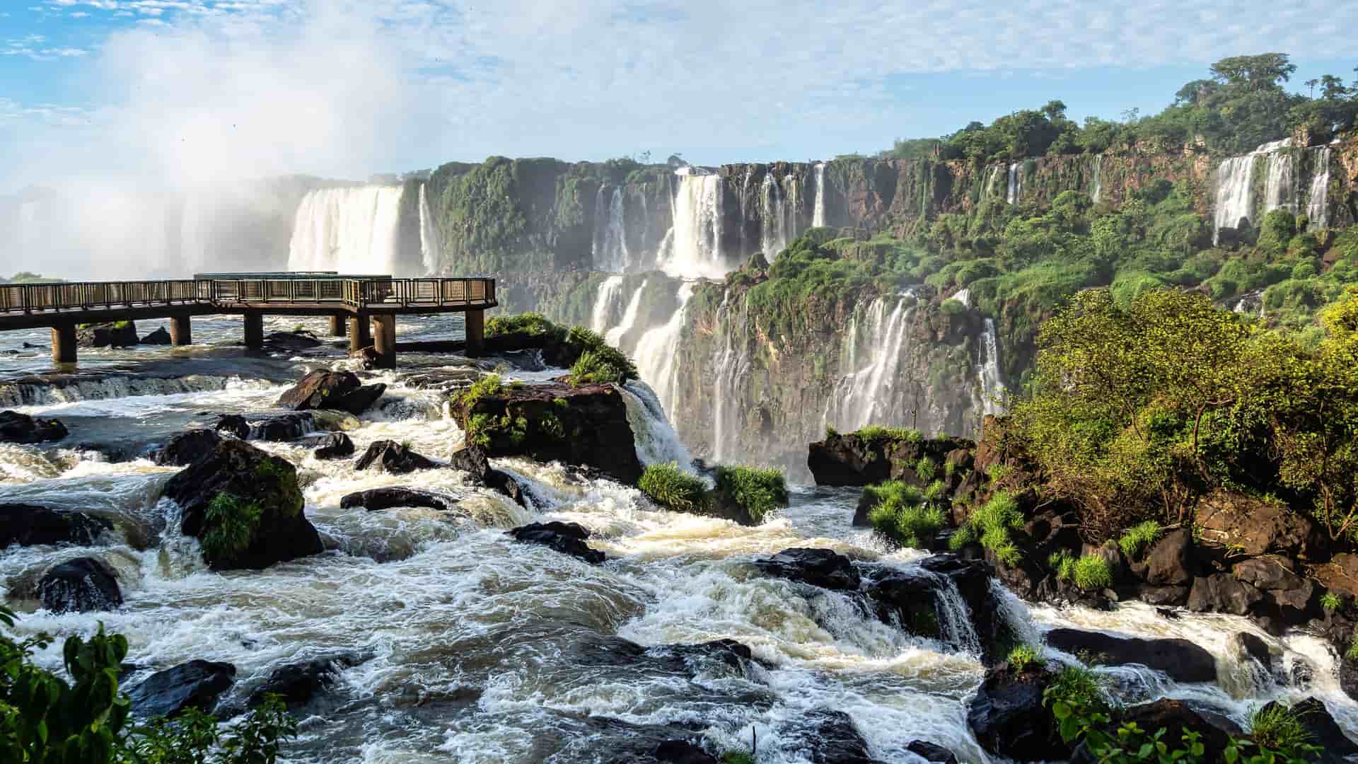 "A beautiful panoramic shot of the stunning Iguazu Falls on the border of Argentina and Brazil, with multiple powerful waterfalls cascading down a cliff face and a wooden walkway stretching over the turbulent river in the foreground. "