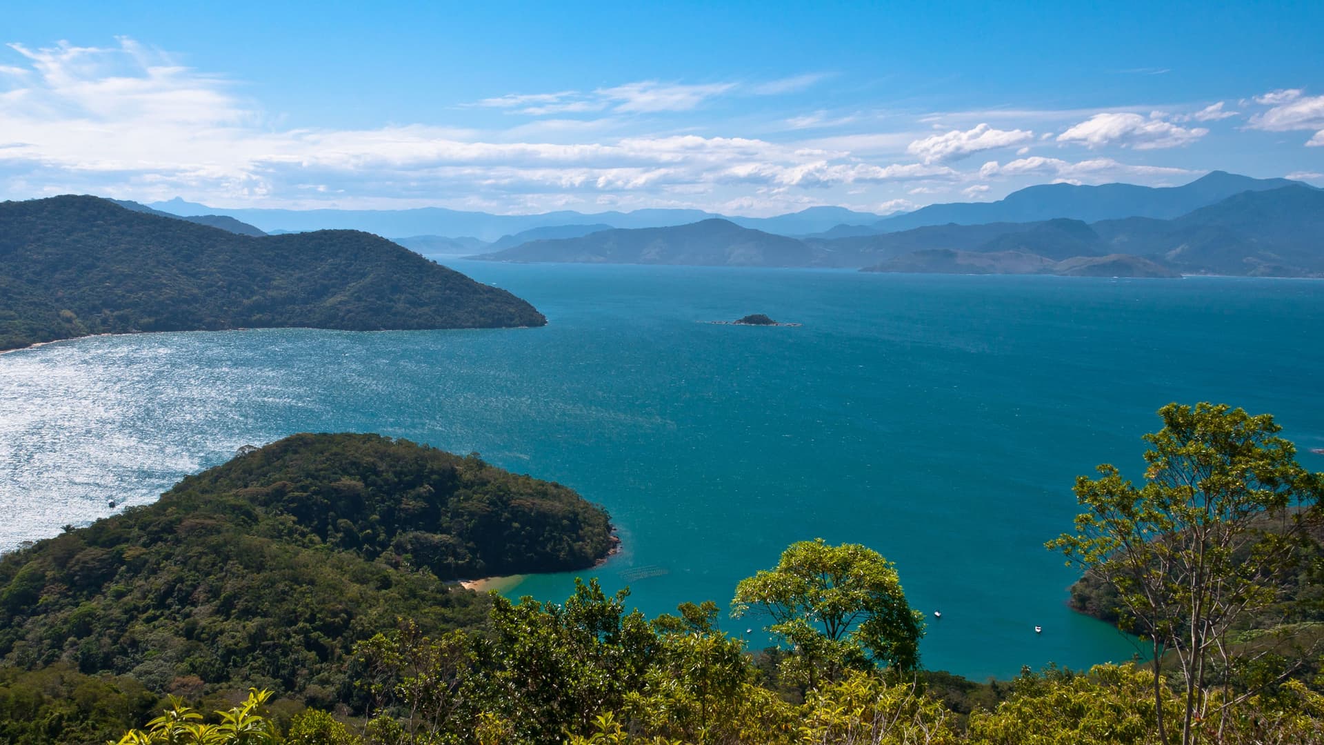 A beautiful panoramic view of the Ilha Grande coastline in Brazil, showcasing the vibrant turquoise sea, lush green tropical islands, and misty mountains on the horizon.