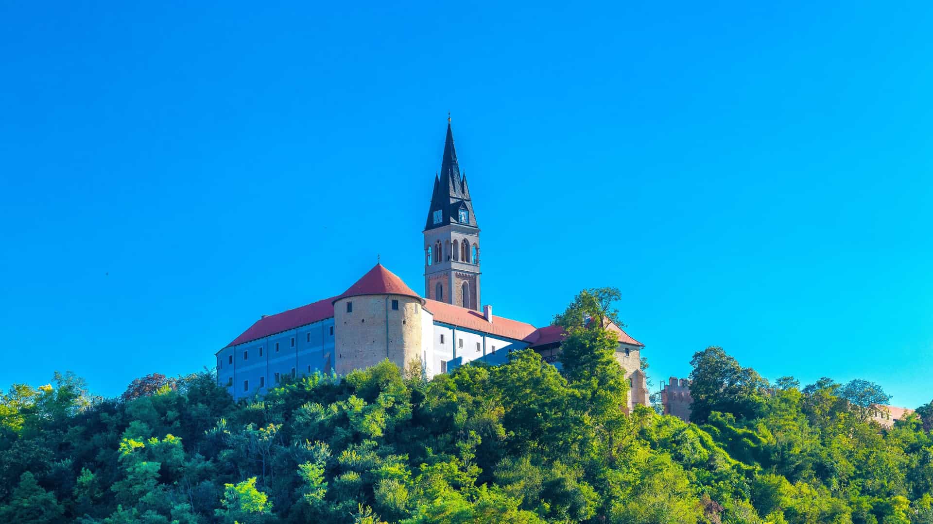 A majestic shot of Ilok Castle in Croatia, showcasing the large fortress with its prominent bell tower and red-roofed buildings perched on a lush, green hill under a clear blue sky.