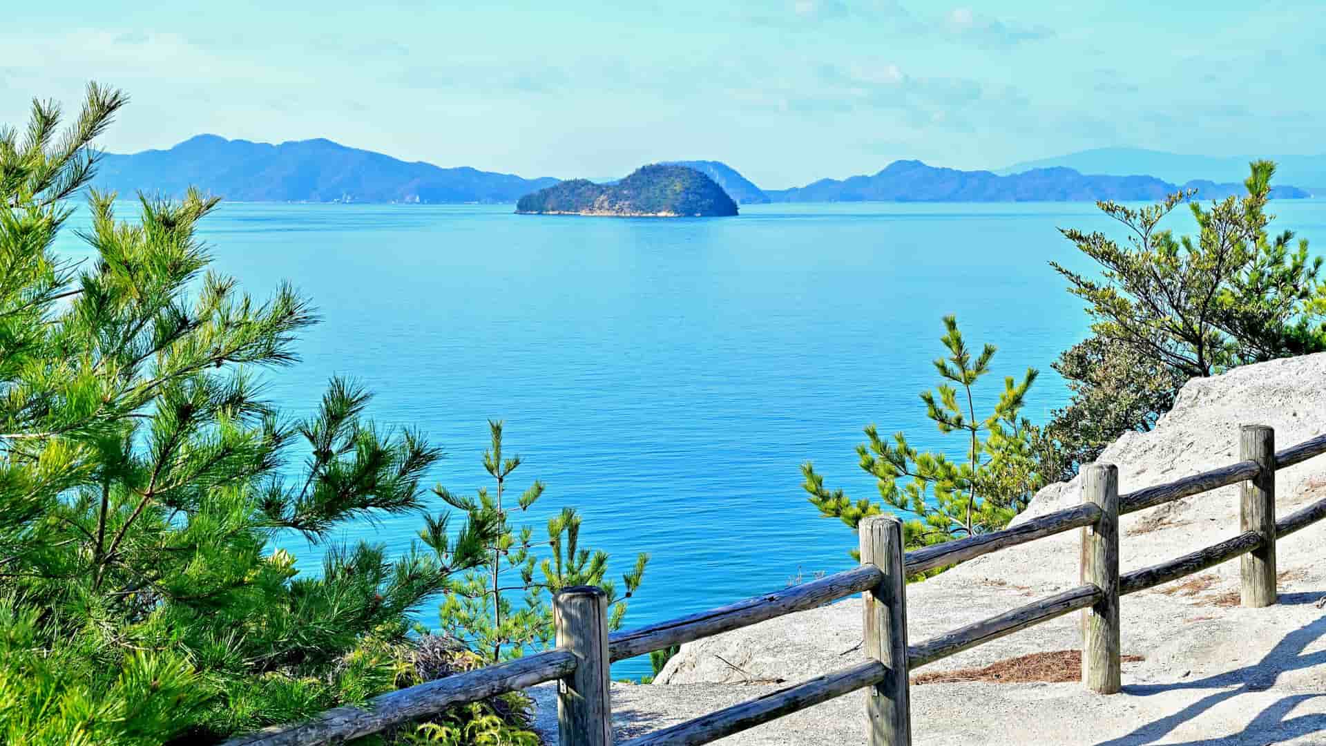 A stunning scenic view of the Inland Sea of Japan, with a tranquil blue body of water, distant islands and mountains, framed by green trees and a wooden fence in the foreground.