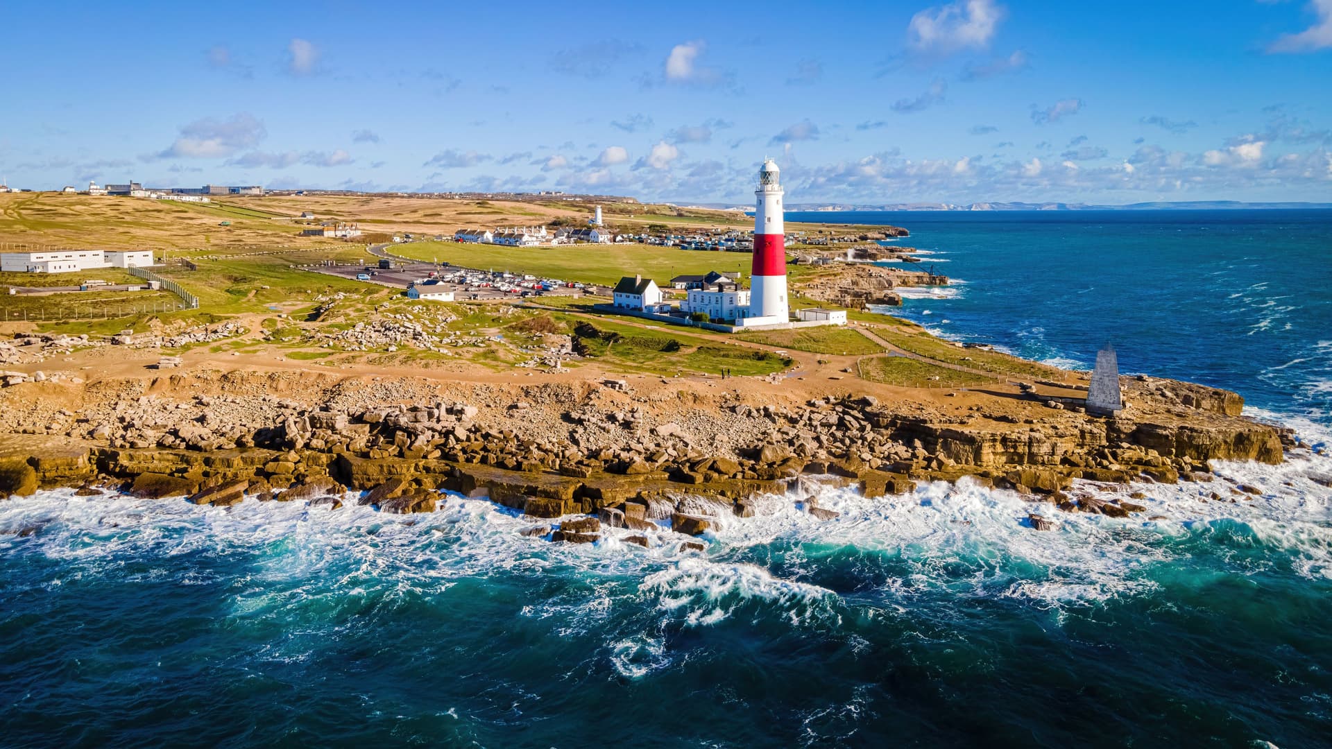 An aerial view of the iconic Portland Bill Lighthouse, with its distinctive red and white stripes, standing on a rocky coastline with waves crashing below.