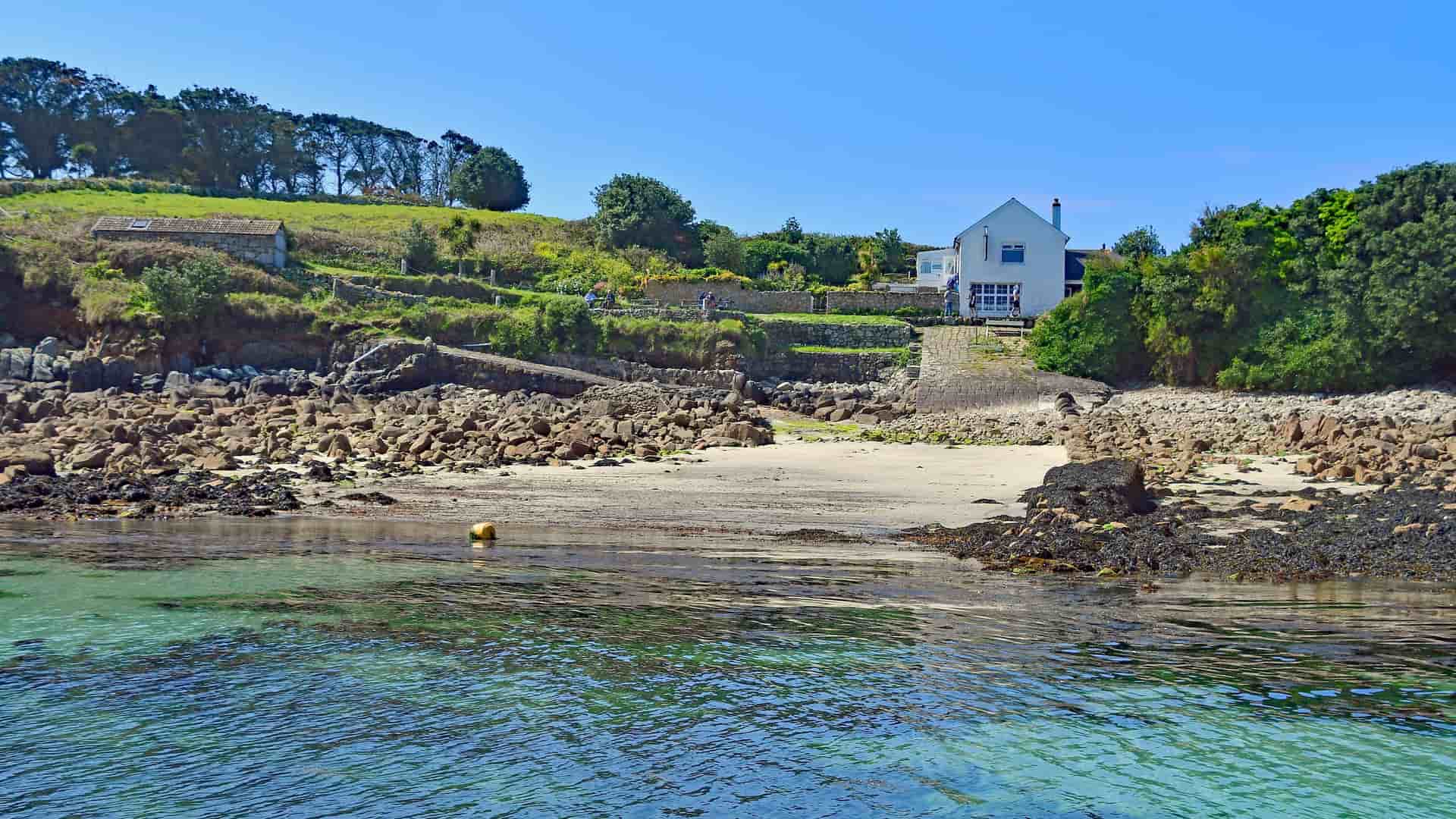 A serene view of a white house with a grassy lawn, situated on a rocky beach with calm turquoise water in the Isle of Scilly.