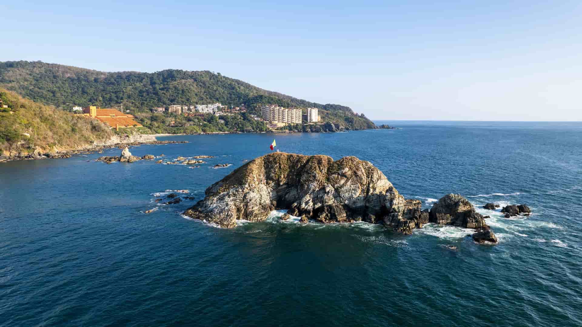 An aerial view of a small rocky island with a Mexican flag on top, off the coast of Ixtapa-Zihuatanejo, with a hillside town and hotels in the background.