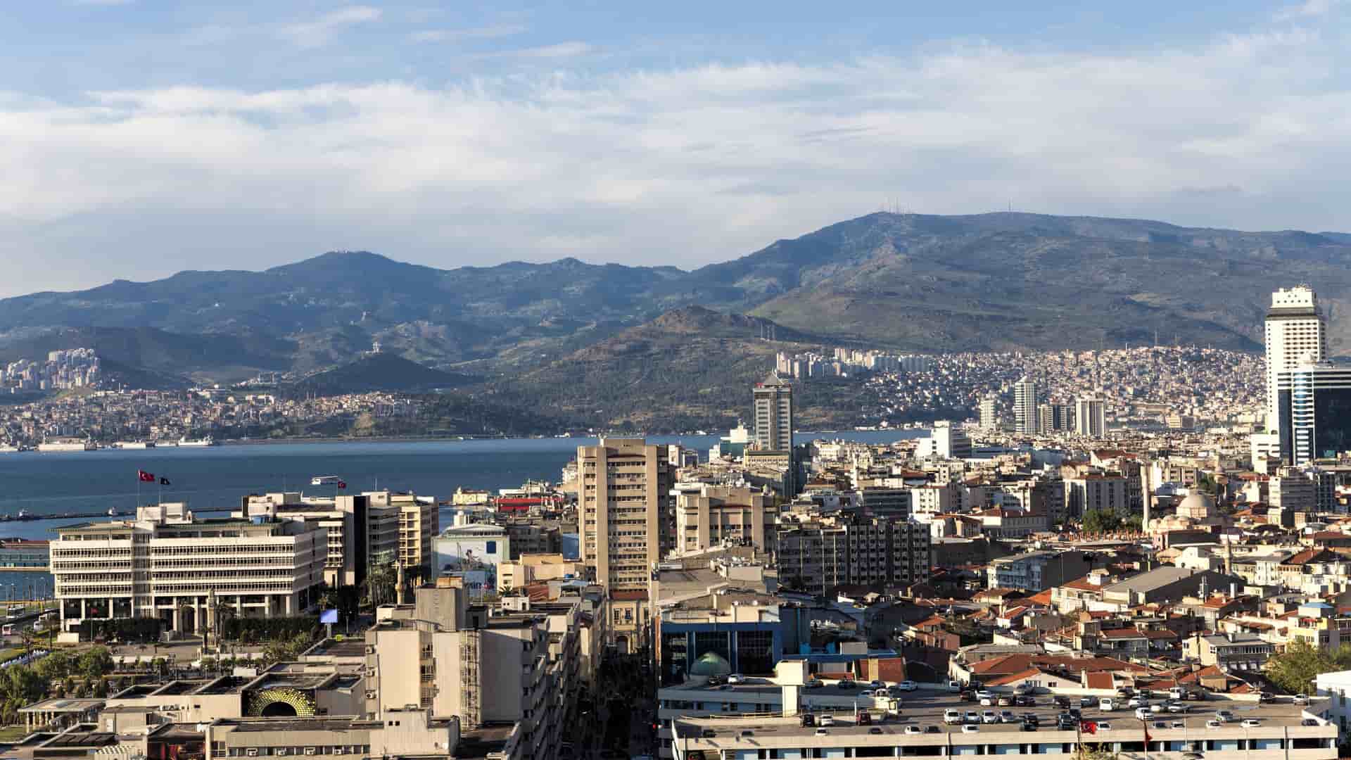 A scenic panoramic view of the bustling city of Izmir, with its modern buildings and historic architecture, nestled between a large bay and a backdrop of mountains.