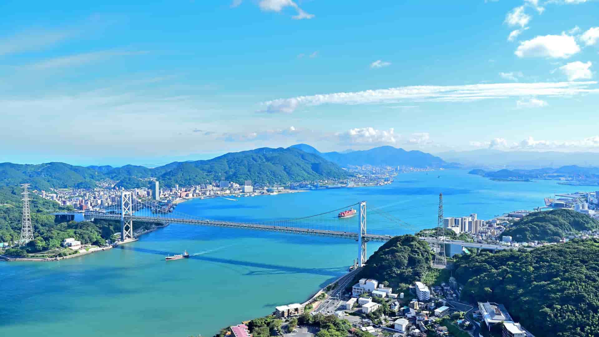 A stunning aerial view of the Kanmon Straits and the Kanmon Bridge, a suspension bridge connecting Honshu and Kyushu, with cityscapes and green mountains in the distance under a bright blue sky.