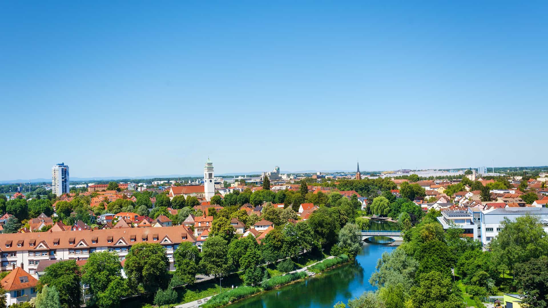 A scenic panoramic view of Kehl, Germany, with a winding river in the foreground, lush green trees, and the city's red-roofed buildings under a clear blue sky.