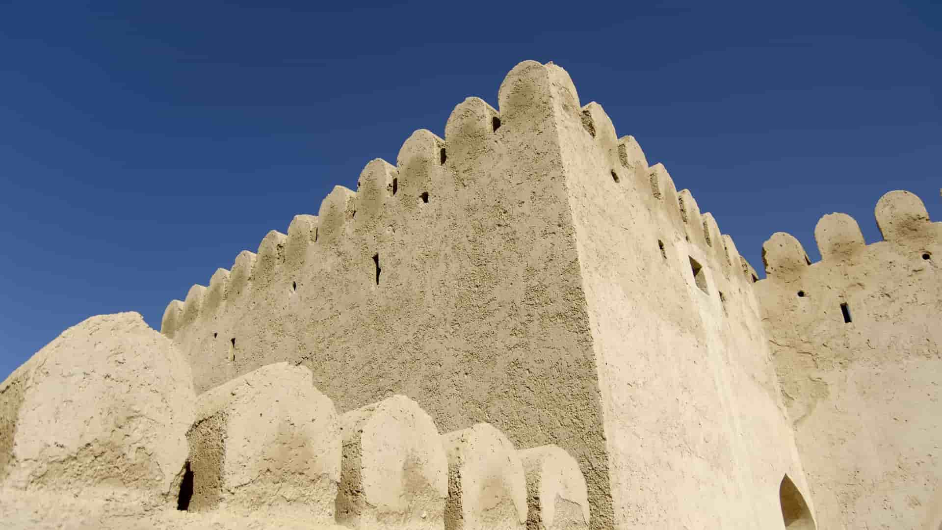 A striking low-angle view of the historic Khasab Fort in Oman, showcasing its crenellated mud-brick walls against a deep blue, cloudless sky.