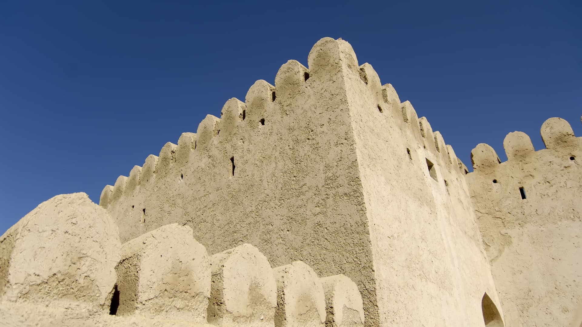 A striking low-angle view of the historic Khasab Fort in Oman, showcasing its crenellated mud-brick walls against a deep blue, cloudless sky.