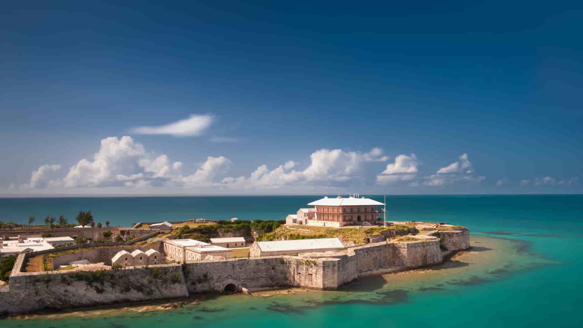 An aerial view of the historic Commissioner's House and the Bermuda National Museum, a white stone fortress on a small island surrounded by stunning turquoise waters near King's Wharf.