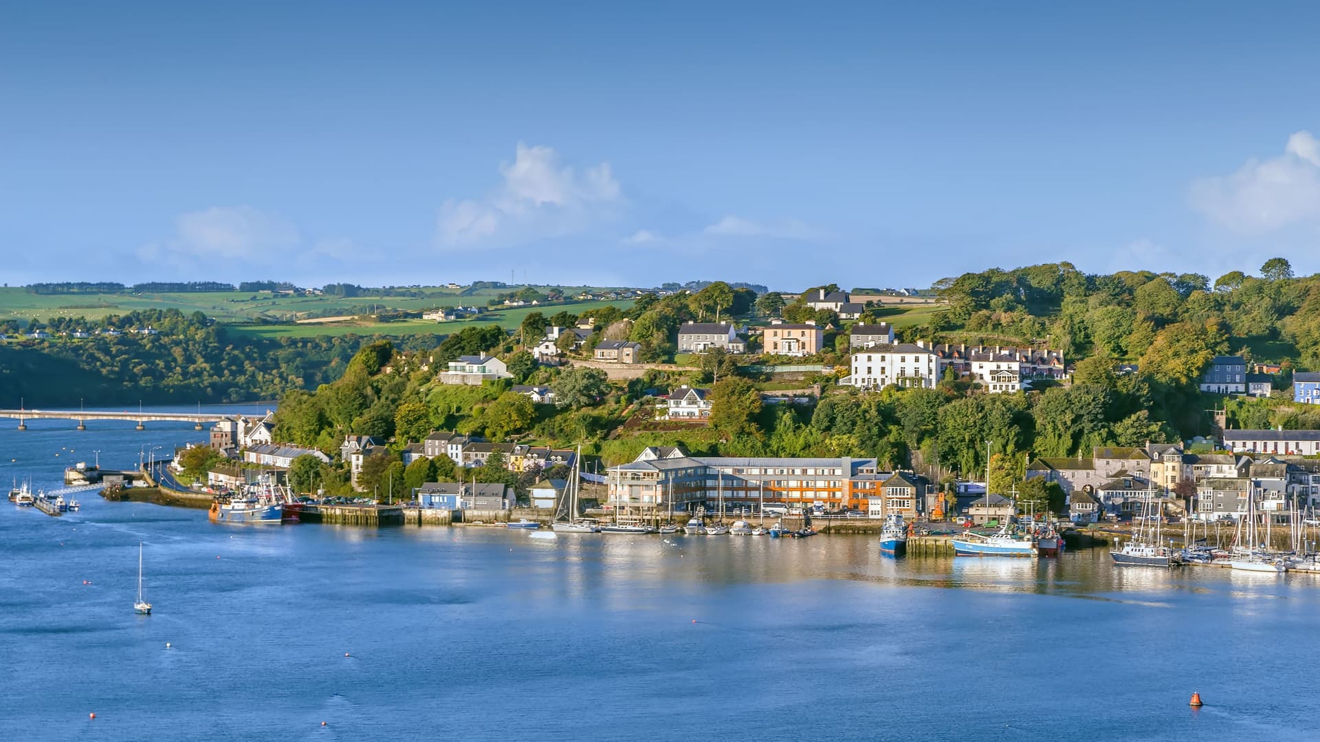 "A beautiful harbor shot of the coastal town of Kinsale, Ireland, with colorful buildings and houses lining the steep hills overlooking the calm water, with boats and yachts moored in the marina. "