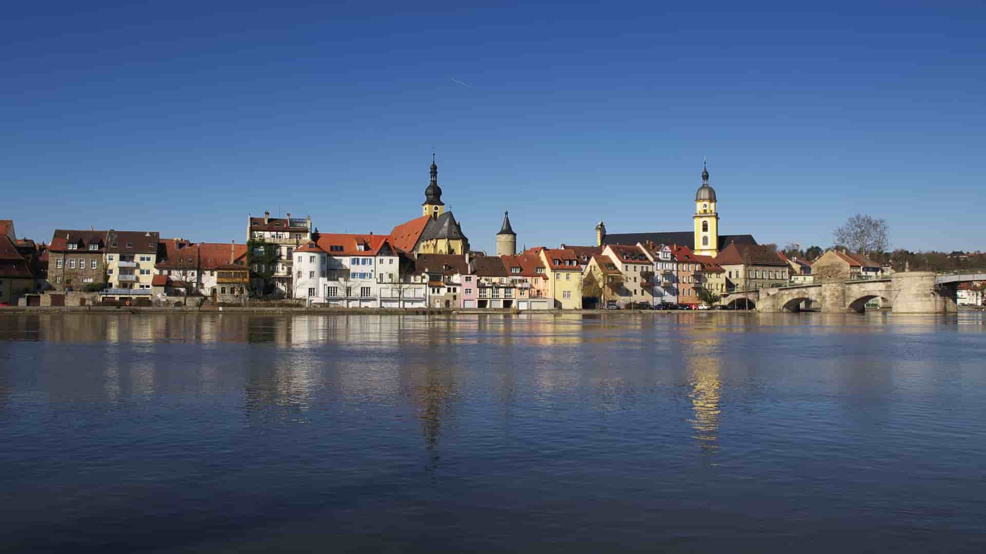 A serene panoramic view of the historic town of Kitzingen, Germany, with colorful buildings and church spires reflecting in the tranquil Main River under a clear blue sky.