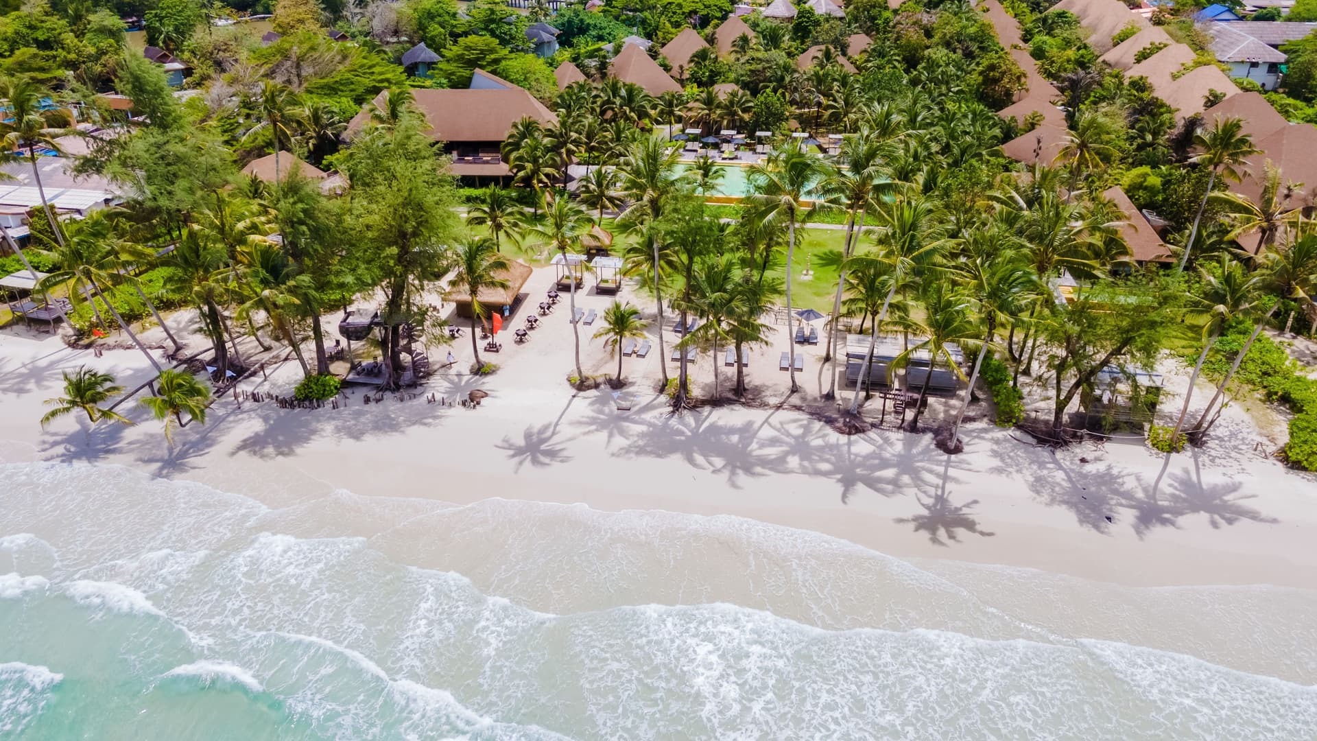 An aerial view of a luxurious resort on Ko Kut (Koh Kood), Thailand, with thatched-roof bungalows, palm trees, and lounge chairs on a white sandy beach with gentle waves.