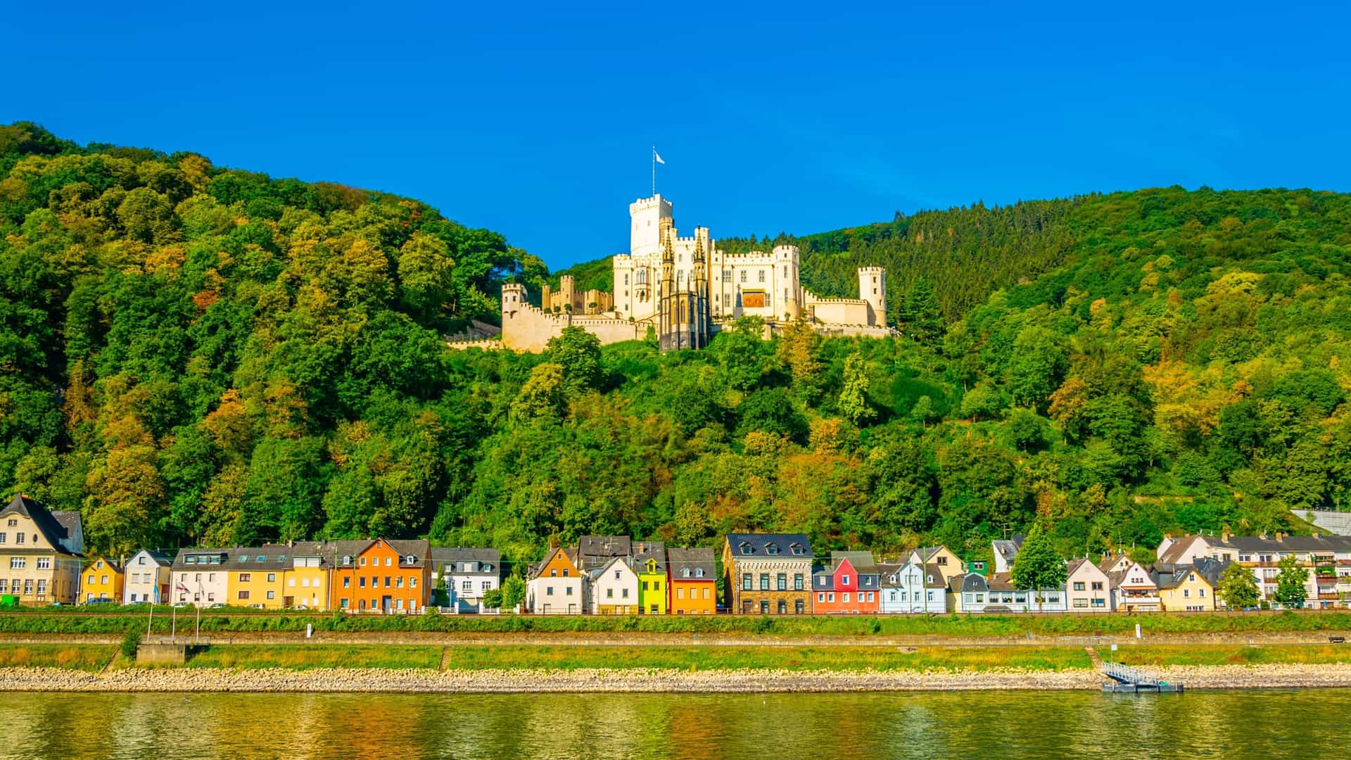 A panoramic view of Koblenz, Germany, with colorful houses lining the riverfront and the impressive Marksburg Castle perched on a lush, forested hill overlooking the town.