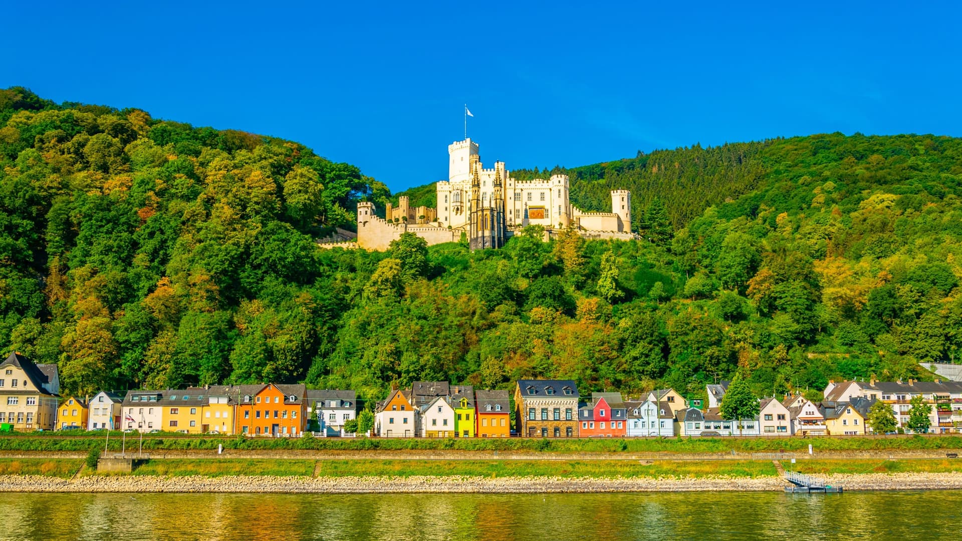 A panoramic view of Koblenz, Germany, with colorful houses lining the riverfront and the impressive Marksburg Castle perched on a lush, forested hill overlooking the town.