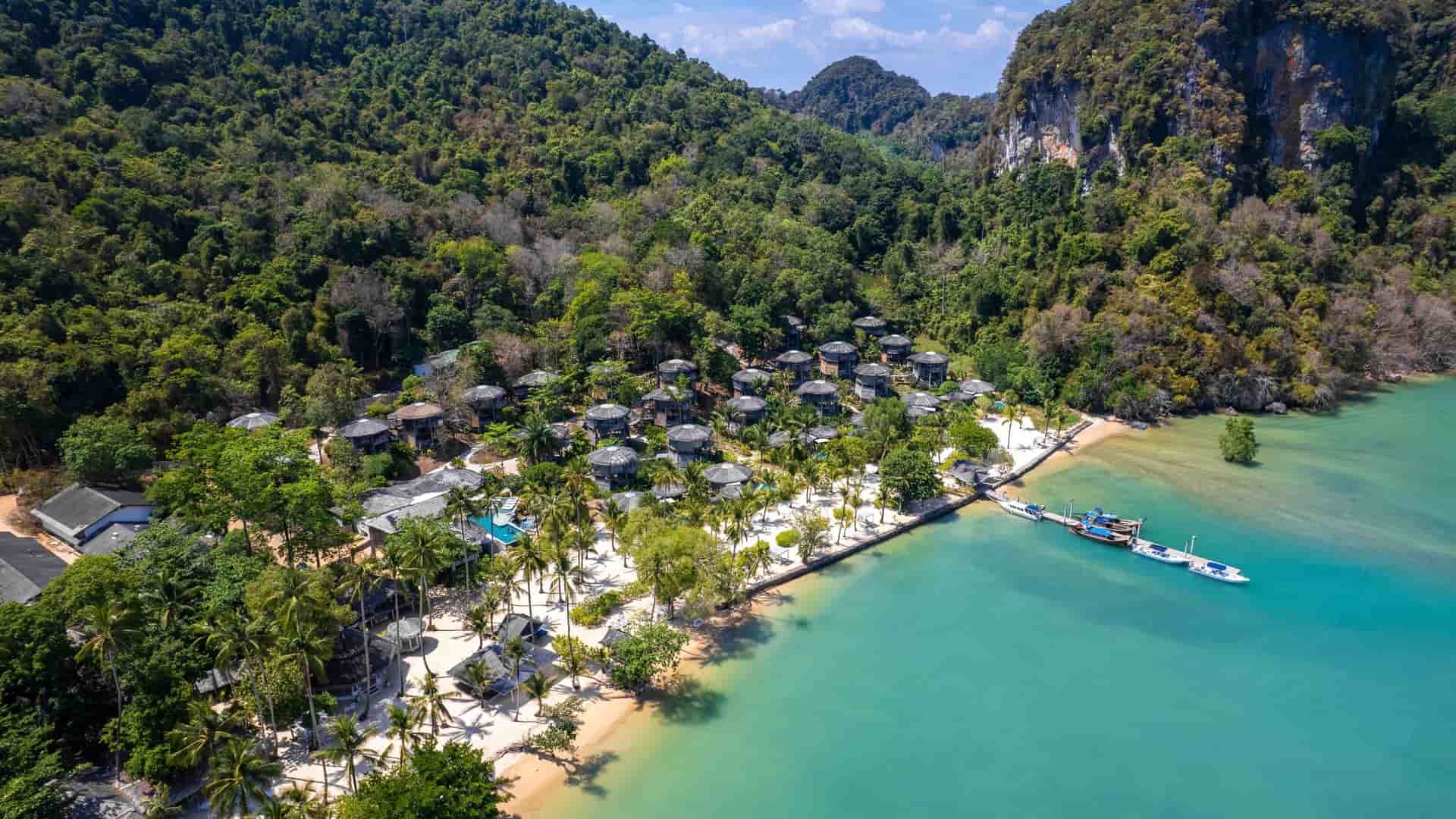 An aerial view of a secluded luxury resort on Koh Yao Noi, Thailand, with bungalows and a swimming pool nestled in lush jungle foliage next to a white sand beach and turquoise water with a boat dock.