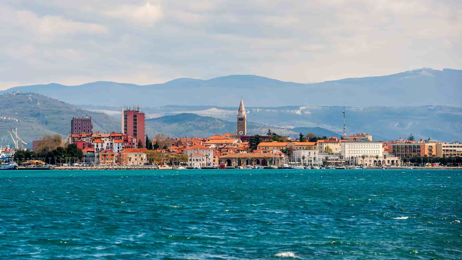 A vibrant panoramic view of Koper, Slovenia, with a beautiful waterfront, historic red-roofed buildings, and a church steeple, set against a backdrop of rolling mountains.