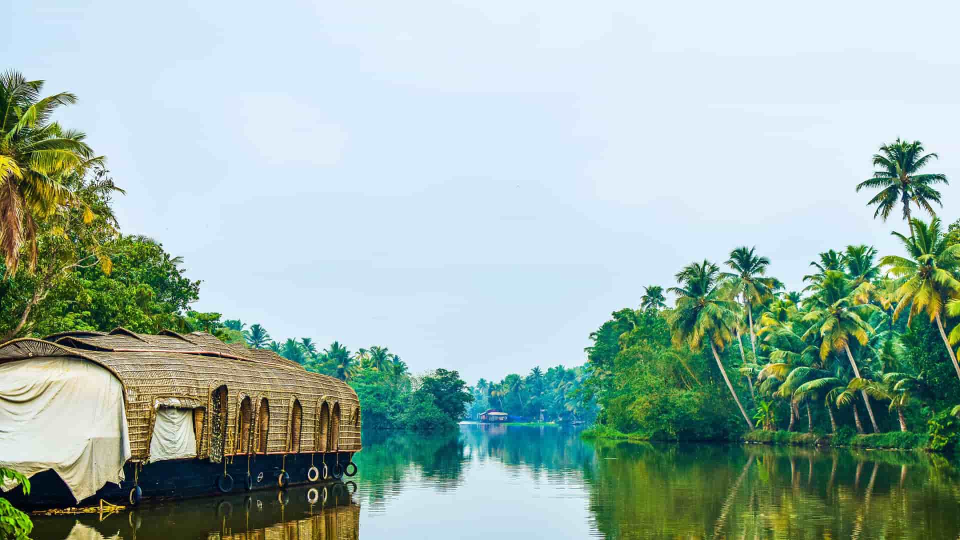 "A scenic view of a traditional houseboat on the tranquil backwaters of Kumarakom, Kerala, India, with lush palm trees and tropical vegetation lining the banks. "