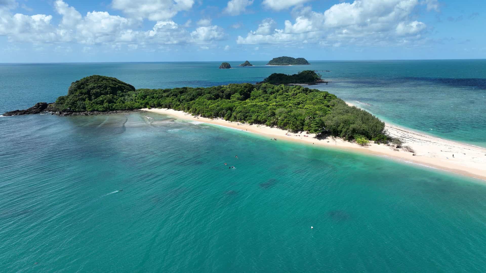 An aerial view of Lady Franklin Island, a remote tropical island with a sandy beach and lush green forest, surrounded by turquoise water under a blue sky with clouds.