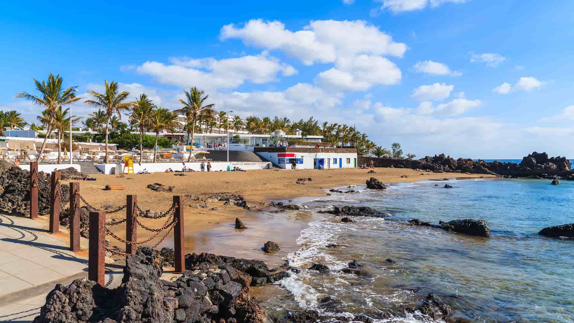 A sun-drenched beach with volcanic rocks and palm trees on the coast of Lanzarote in the Canary Islands, Spain, with a resort hotel and a small beach bar visible in the background.