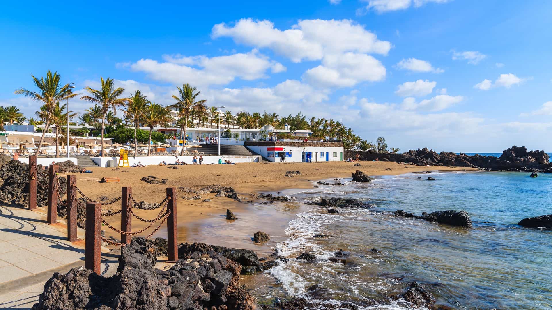 A sun-drenched beach with volcanic rocks and palm trees on the coast of Lanzarote in the Canary Islands, Spain, with a resort hotel and a small beach bar visible in the background.