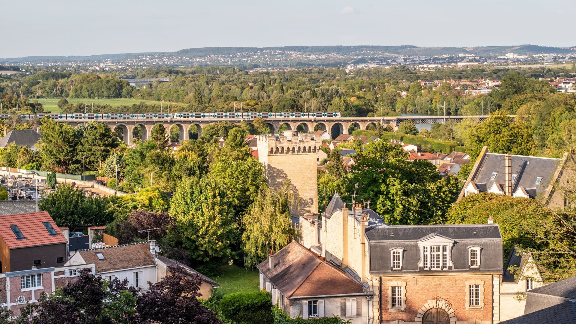 A high-angle view of the charming French town of Le Pecq, showcasing traditional European rooftops and a historic aqueduct carrying a modern train across the Seine River valley.