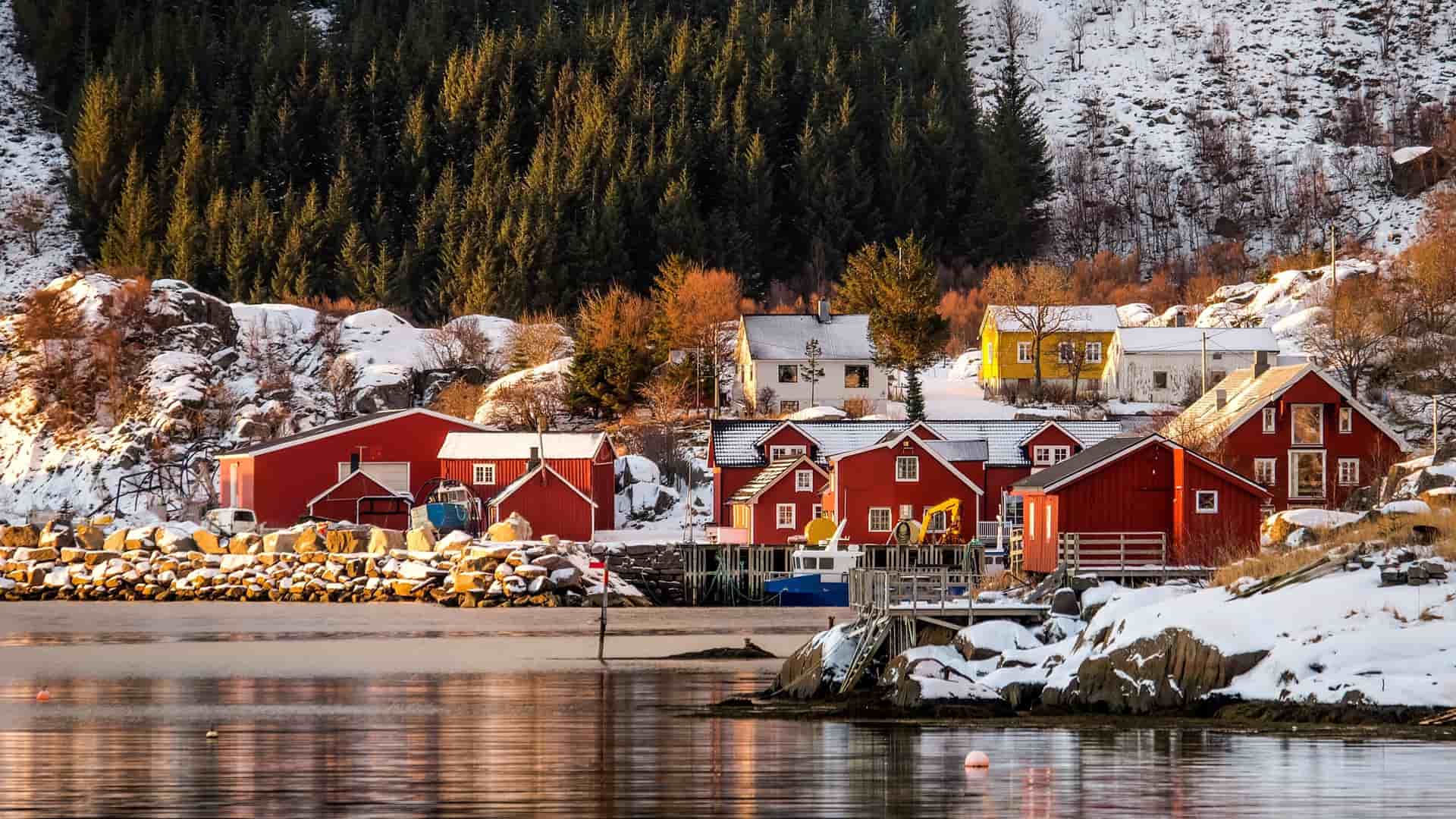 A snowy winter landscape of Leknes, Norway, showing traditional red rorbu cabins and white houses on the shore of a calm harbor with a dense forest and snow-covered hills in the background.