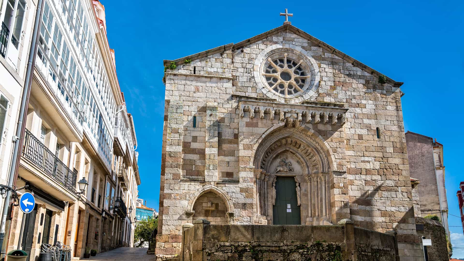 "A low-angle view of the historic Romanesque Church of Santa Maria de Fisterra in Lestedo, Spain, featuring its stone facade, arched entrance, and a rose window. "