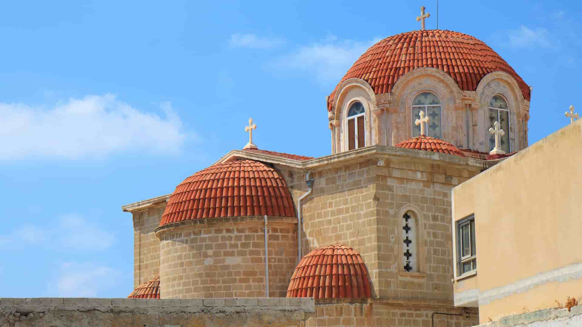 A detailed shot of a traditional, stone-built Greek Orthodox church in Limassol, Cyprus, with multiple red-tiled domes and golden crosses against a blue sky.