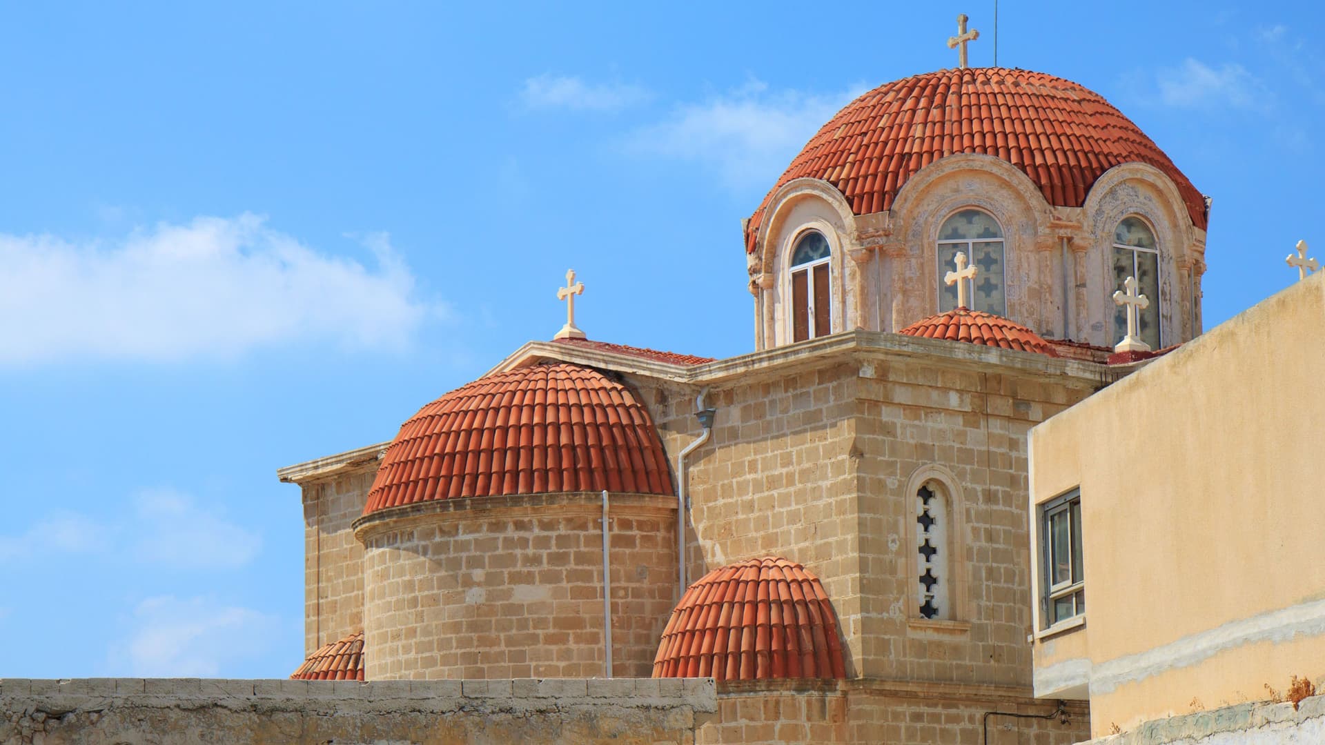 A detailed shot of a traditional, stone-built Greek Orthodox church in Limassol, Cyprus, with multiple red-tiled domes and golden crosses against a blue sky.