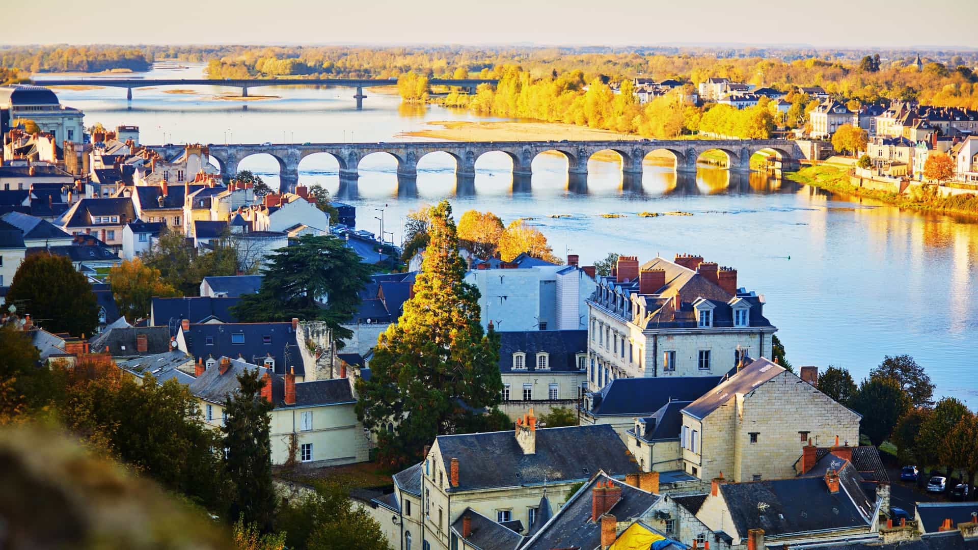 A scenic view of the Loire River in Saumur, France. The Pont Cessart (Cessart Bridge), a long, historic masonry arched bridge, stretches across the wide, shallow river. In the background, the town of Saumur is visible, with the prominent white structure of the Château de Saumur rising above the buildings on the riverbank.
