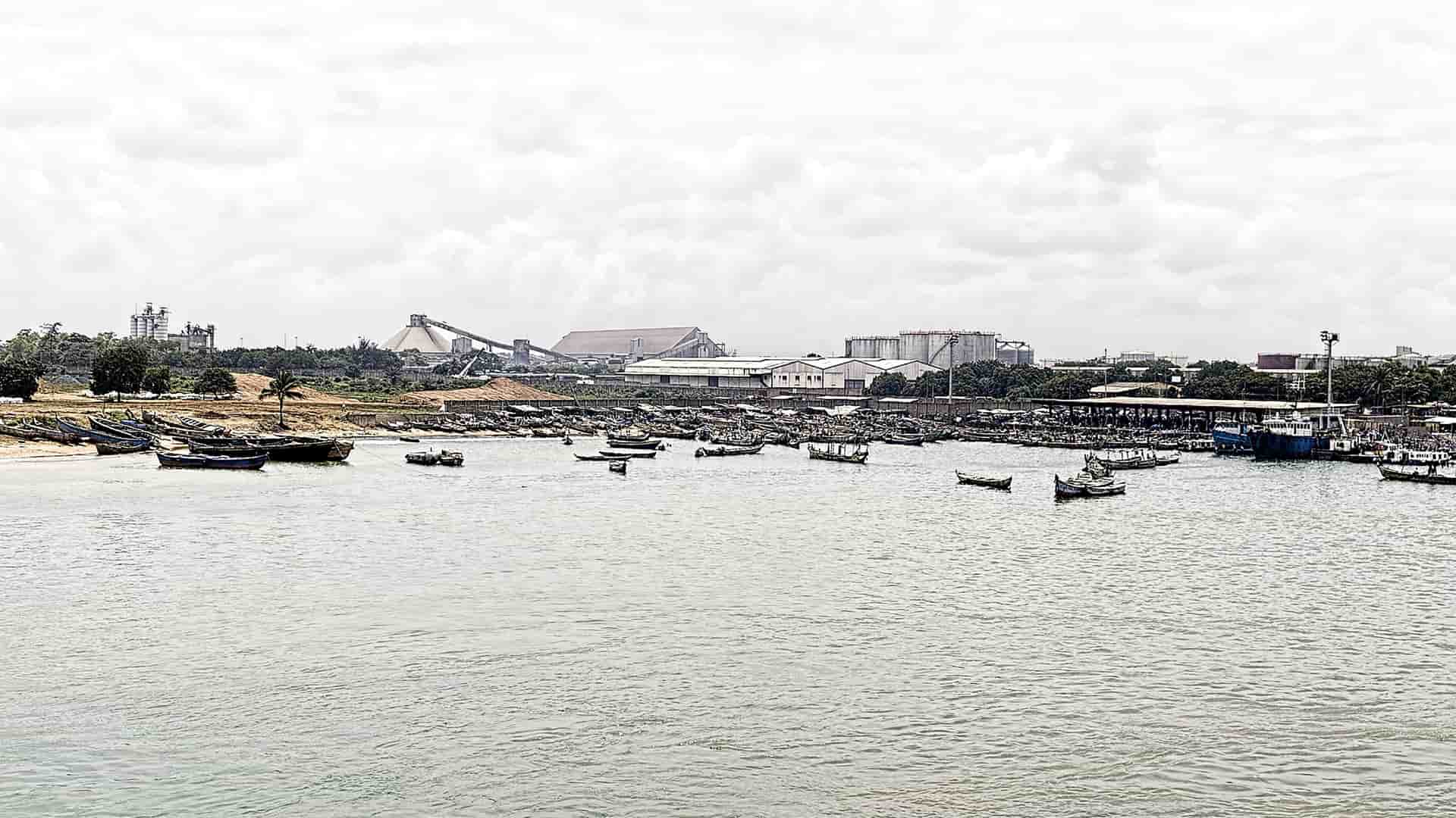 A busy and wide view of the LomƩ seaport in Togo, West Africa, with numerous small fishing boats in the harbor and industrial buildings along the shoreline.