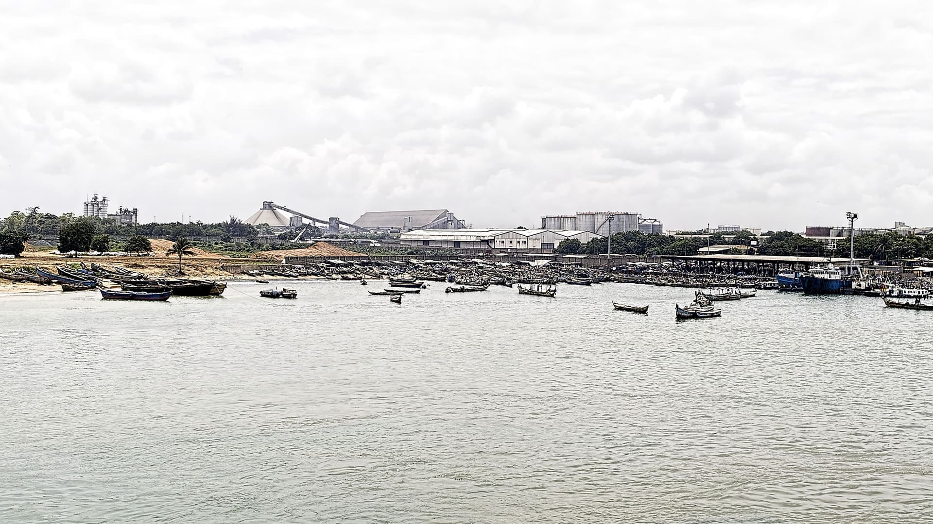 A busy and wide view of the Lomé seaport in Togo, West Africa, with numerous small fishing boats in the harbor and industrial buildings along the shoreline.