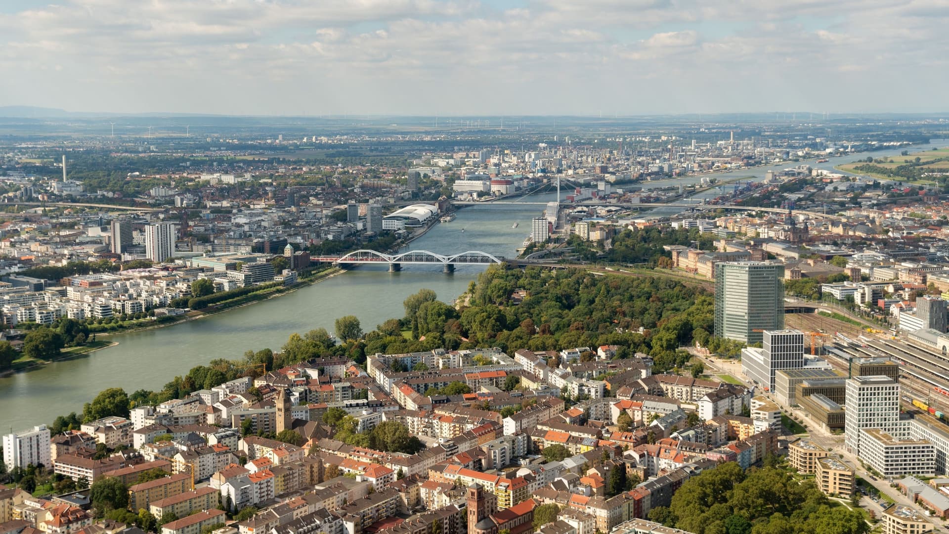 An aerial panoramic shot of Ludwigshafen and Mannheim in Germany, showcasing the Rhine River flowing through the cities, with bridges and modern high-rises.