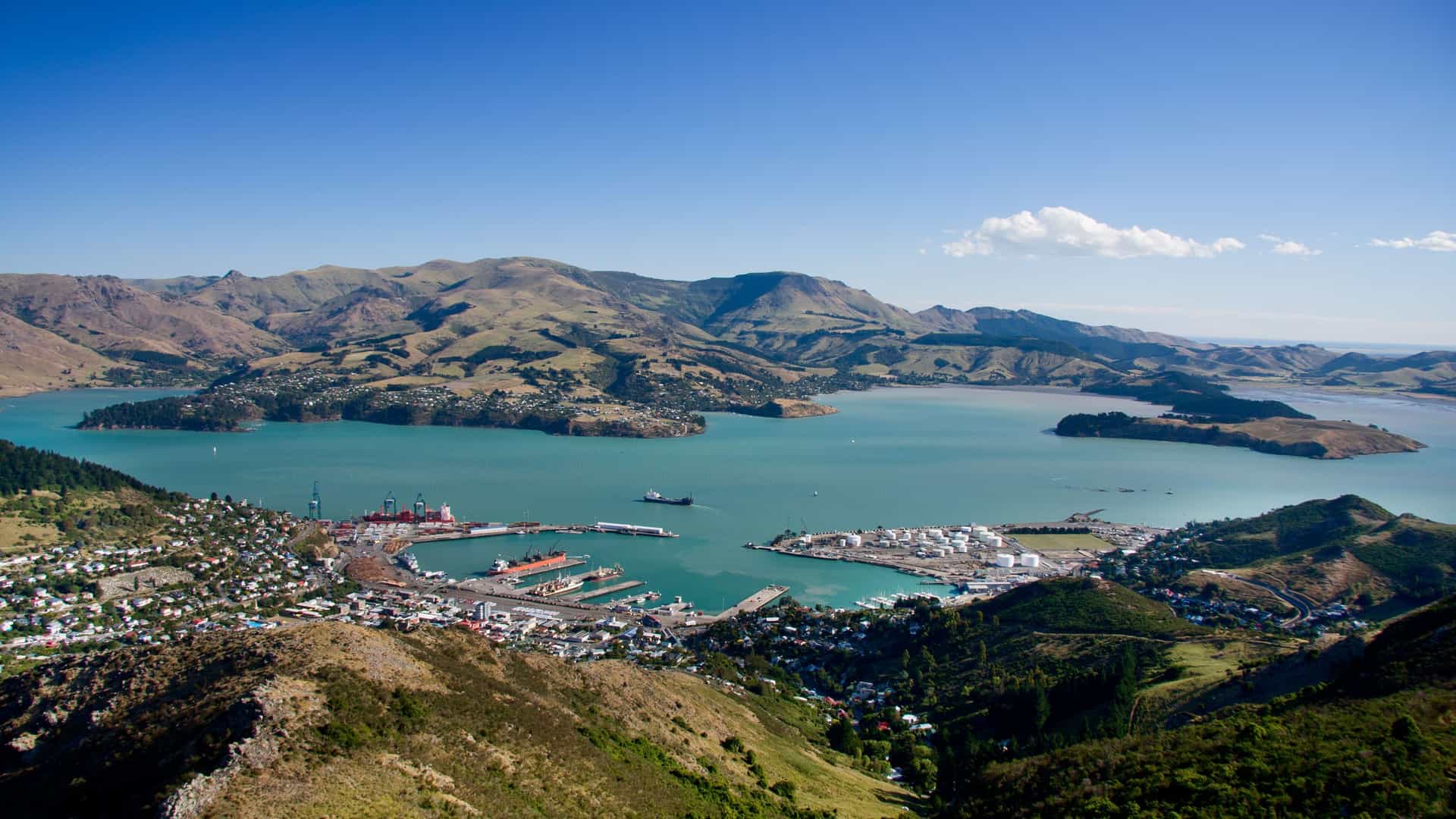 An aerial panoramic shot of Lyttelton Harbour in Christchurch, New Zealand, showcasing the busy port town nestled between the sea and the Port Hills on a clear, sunny day.