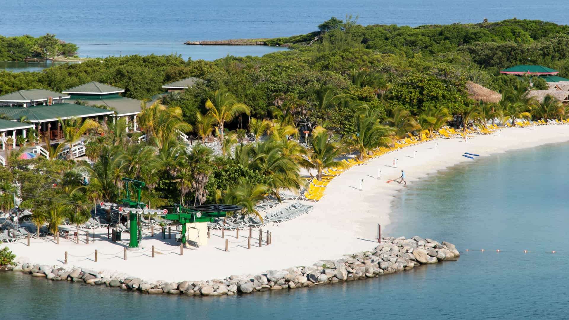 A sunny shot of Mahogany Bay, Roatan, with white sandy beaches, green palm trees, a cruise ship port, and a chairlift.