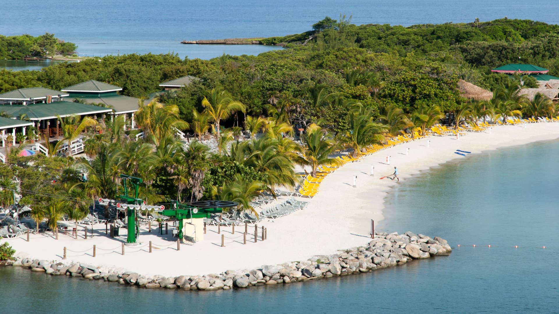 A sunny shot of Mahogany Bay, Roatan, with white sandy beaches, green palm trees, a cruise ship port, and a chairlift.