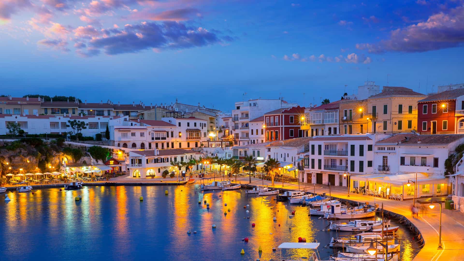 A vibrant waterfront shot of Mahón, Menorca, with boats moored in the harbor and illuminated buildings along the port at dusk.