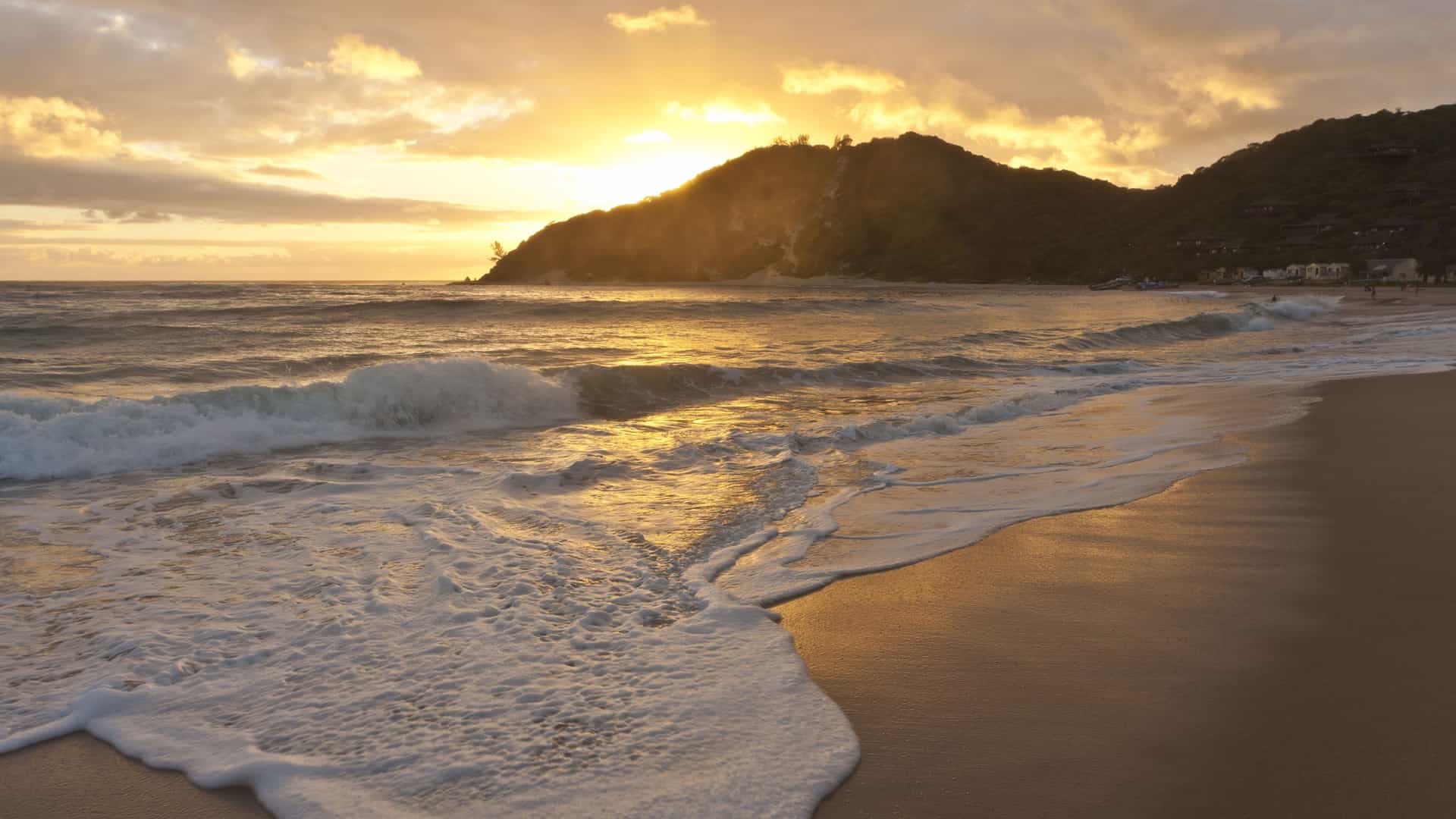 A golden sunset over the Indian Ocean with waves crashing onto a sandy beach in Maputo, Mozambique, with a distant hill silhouetted against the sky.