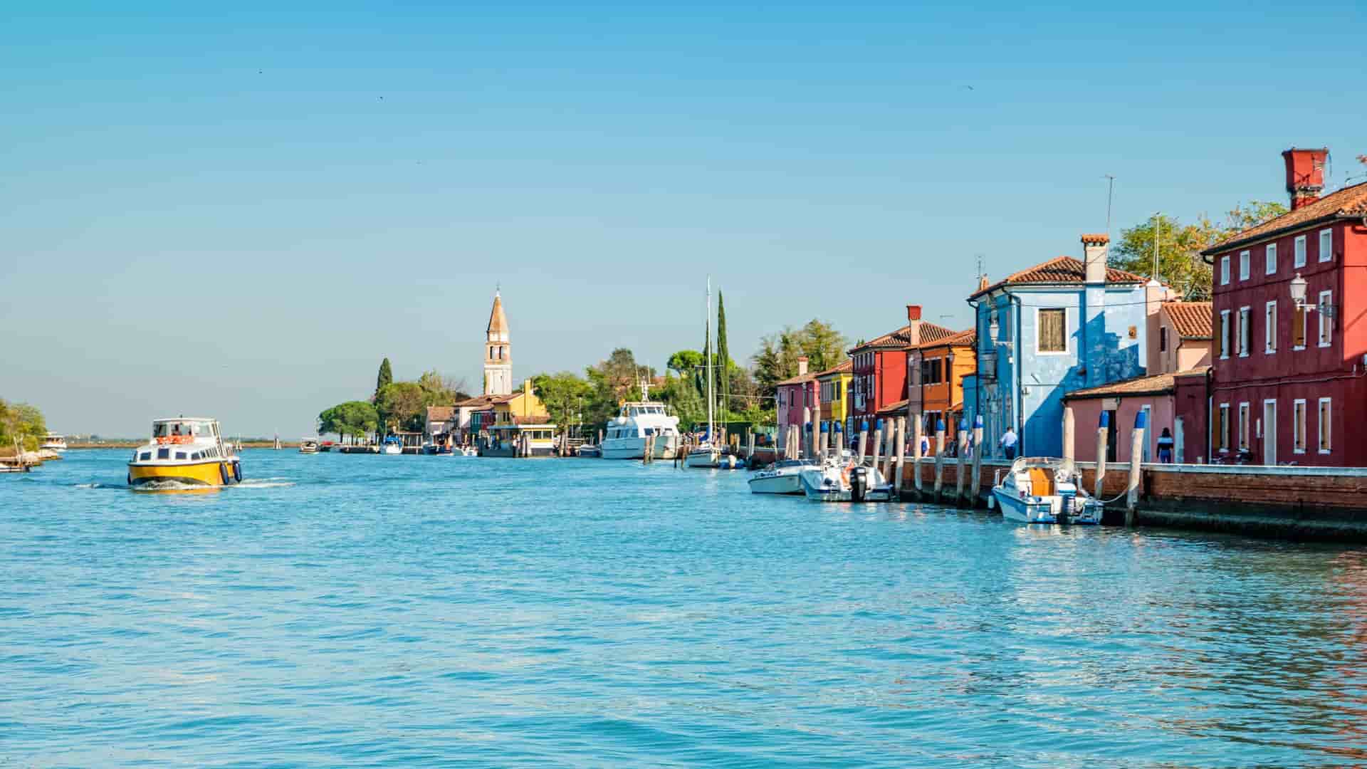 A beautiful canal view on Mazzorbo Island, with colorful buildings lining the water, docked boats, and a church tower visible in the distance.