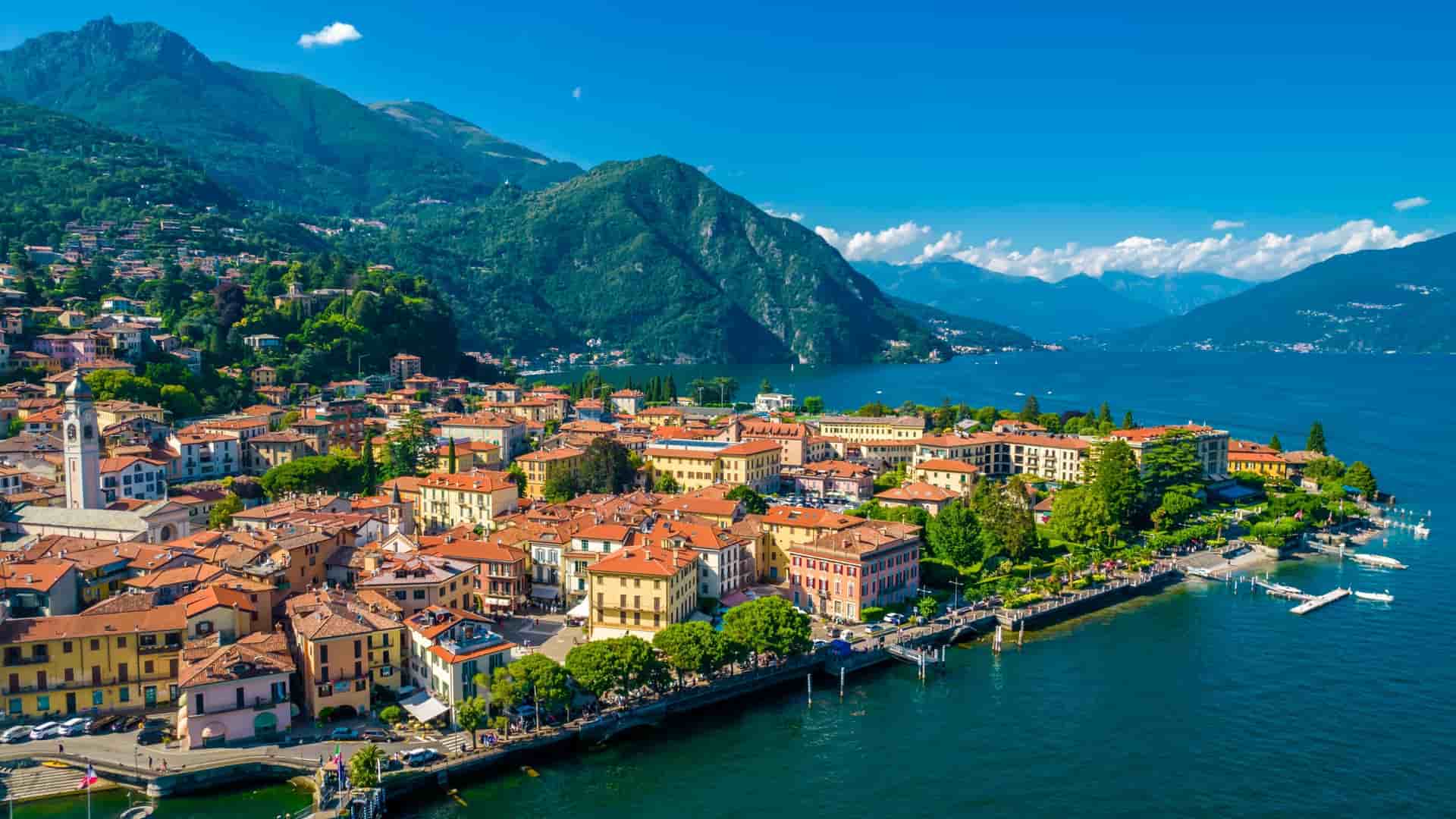 An aerial view of the lakeside town of Menaggio, Italy, with red-roofed buildings nestled against steep green mountains.