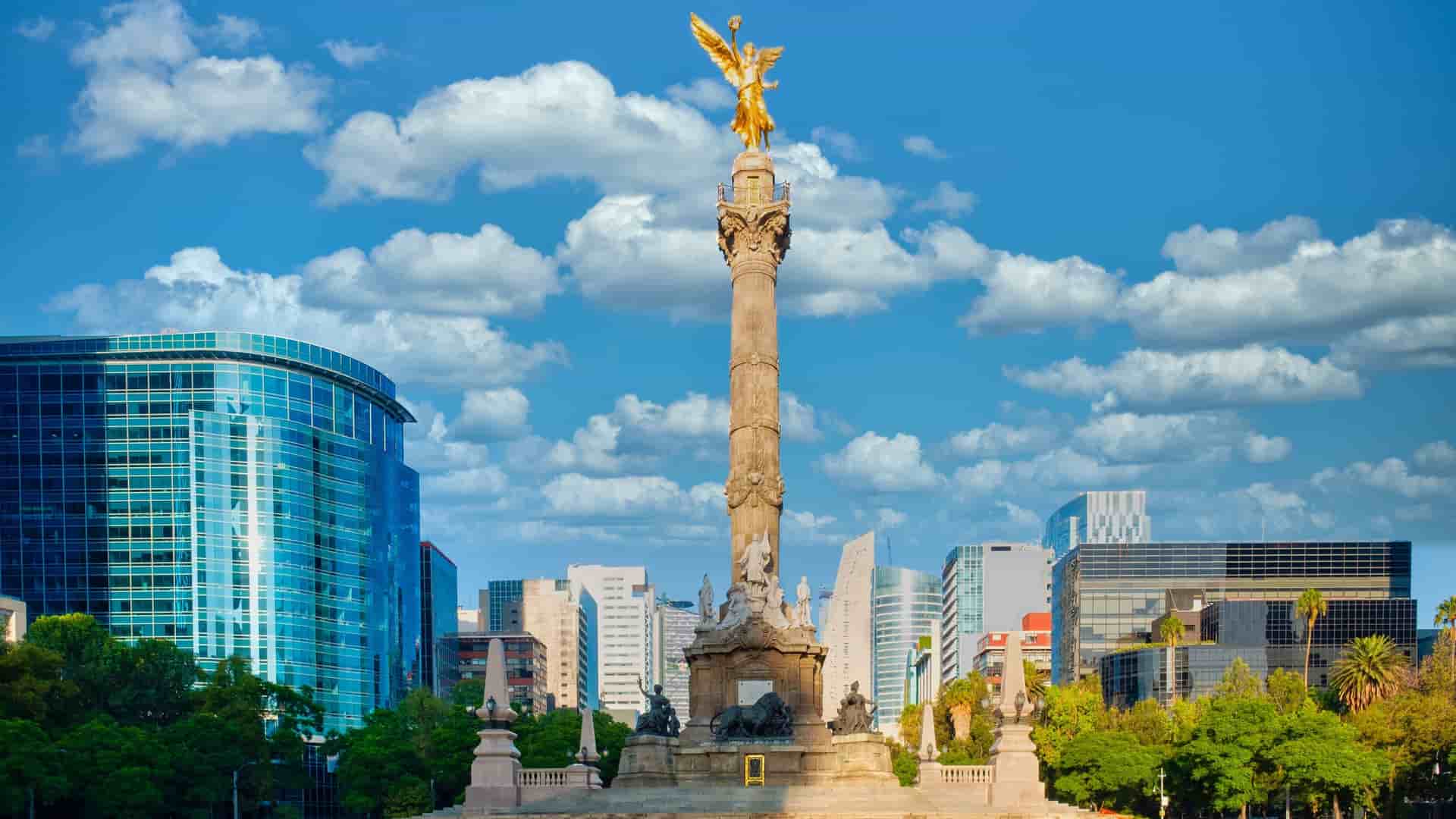 The golden Angel of Independence monument (El Ángel de la Independencia) standing tall against a bright blue sky with modern glass skyscrapers in the background on Paseo de la Reforma in Mexico City.