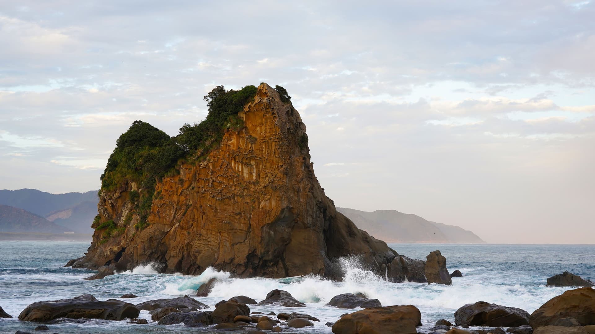 A massive, rugged sea stack with trees growing on its top, located in the coastal waters of Aburatsu, Miyazaki, Japan, with waves crashing against its rocky base.