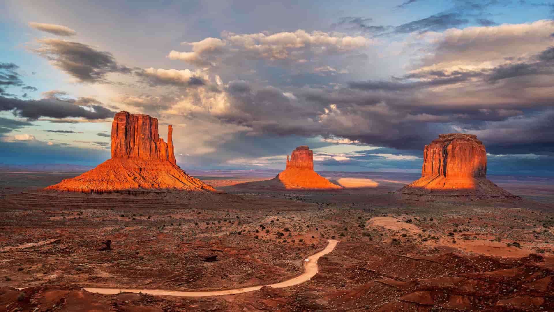 "A dramatic panoramic view of the iconic sandstone buttes and mesas in Monument Valley, Utah, glowing orange at sunset under a moody, cloudy sky. "