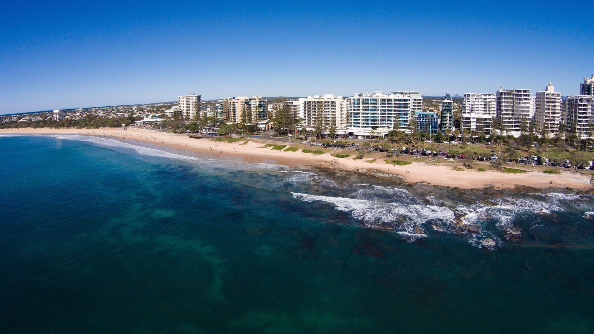 An aerial view of the sunny Mooloolaba Beach, Australia, showcasing a long stretch of golden sand, a calm blue ocean, and a line of tall resort buildings on the coastline.