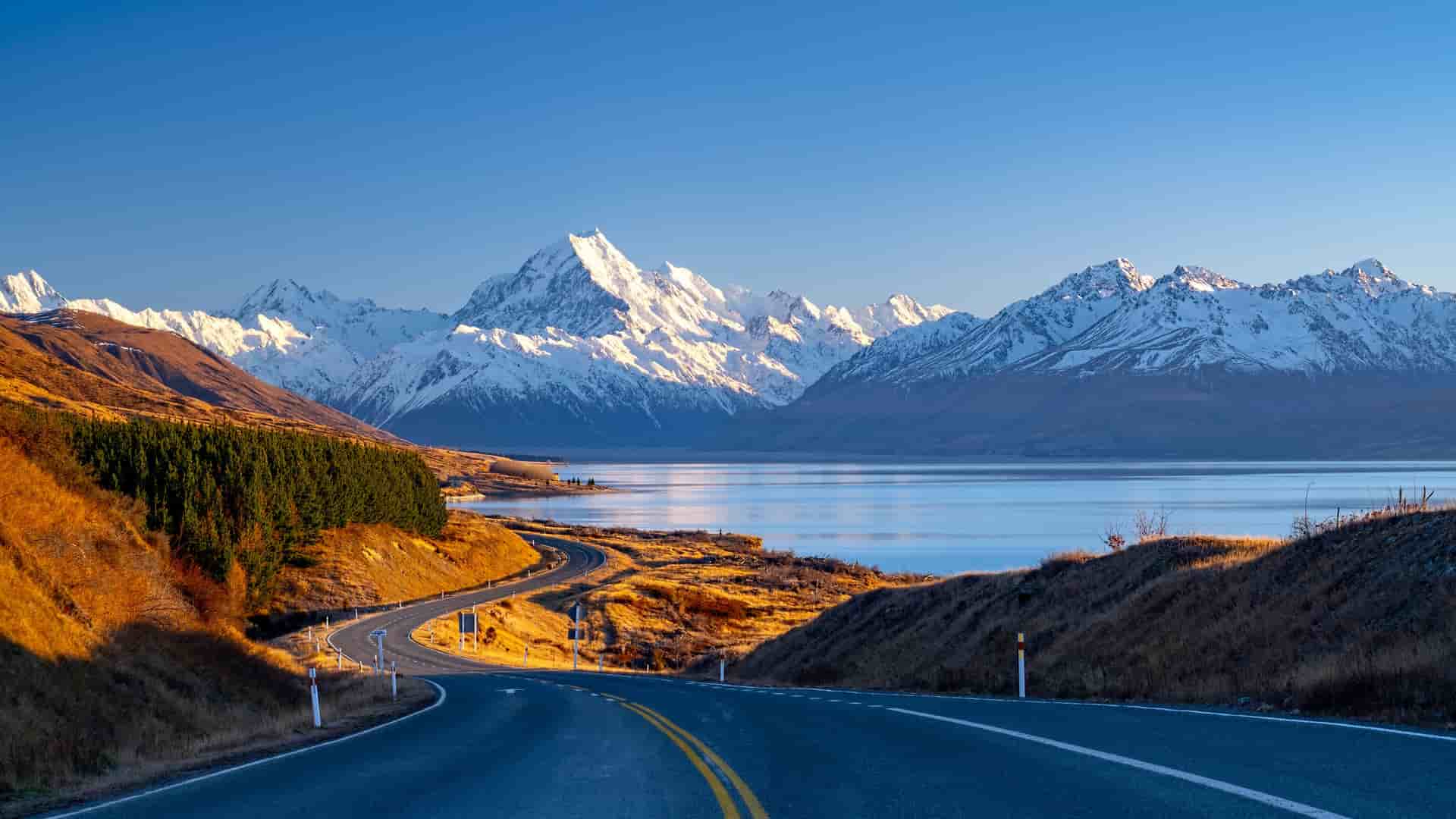 "A beautiful panoramic shot of Mount Cook National Park in New Zealand, with the iconic snow-capped Aoraki Mount Cook mountain range overlooking the bright blue waters of Lake Pukaki at sunrise. "