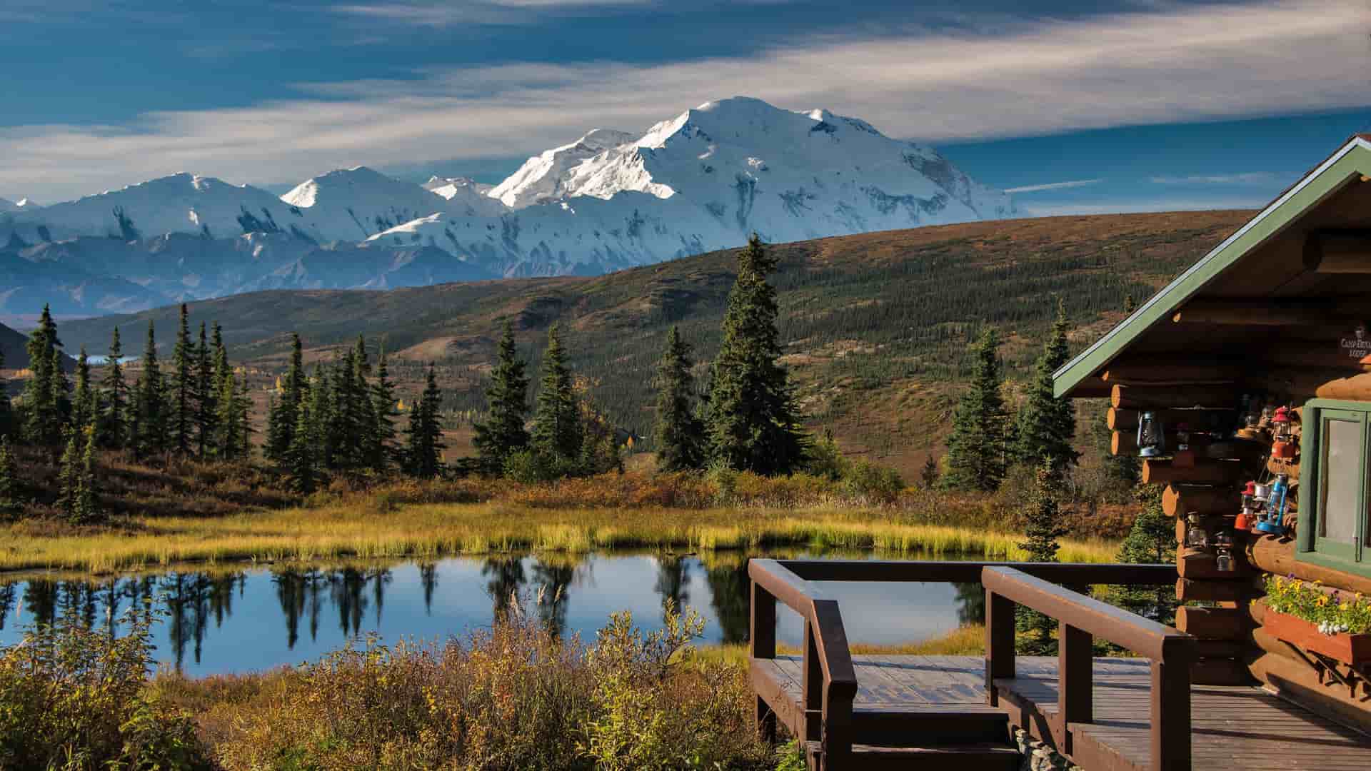 A rustic log cabin deck overlooking a small pond with the majestic, snow-covered Denali mountain range in the background at the Mt. McKinley Princess Wilderness Lodge.