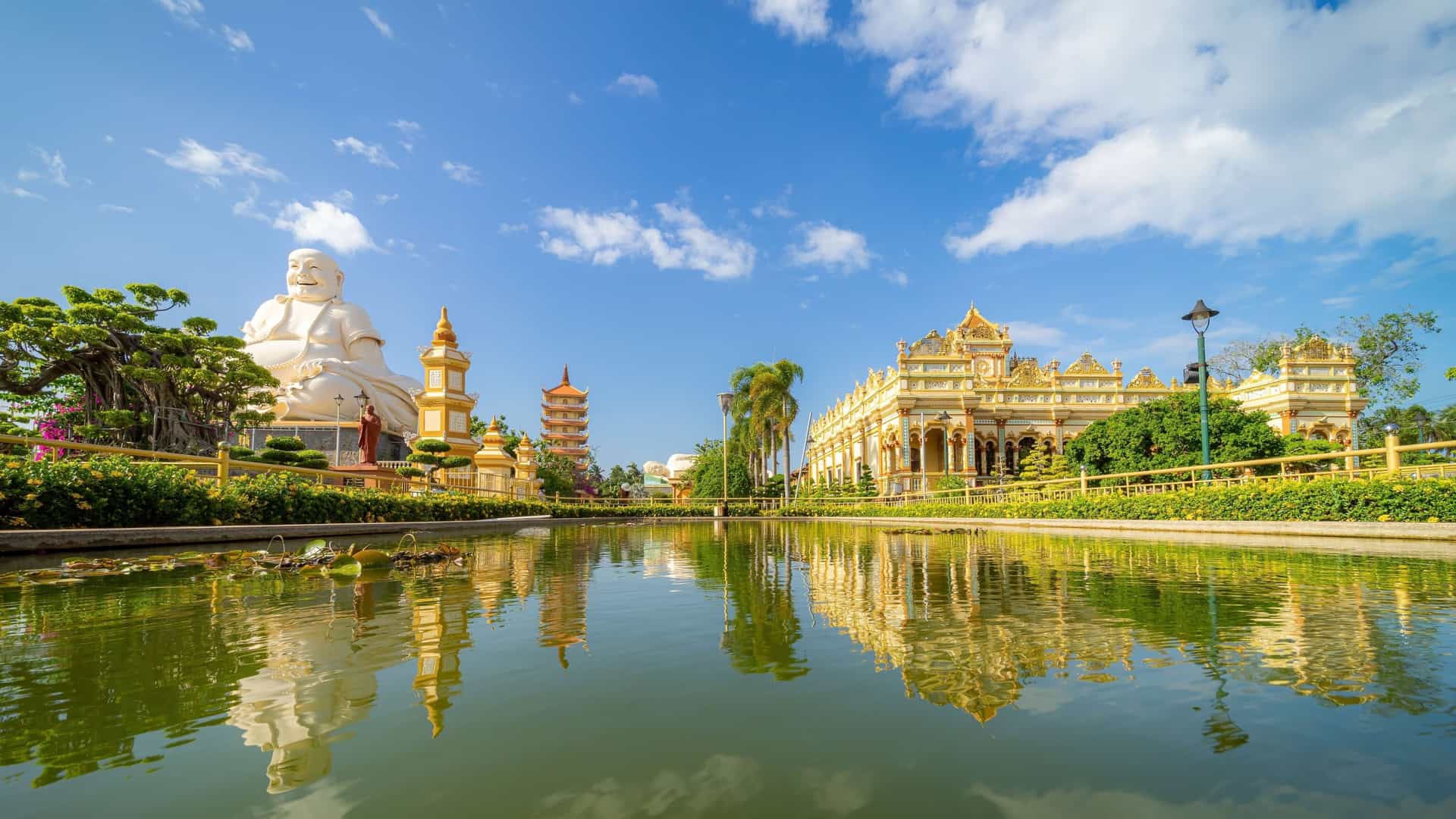 A beautiful Buddhist temple complex in My Tho, Vietnam, with a large, laughing Buddha statue, a pagoda, and a decorative main building reflected in a serene pond.